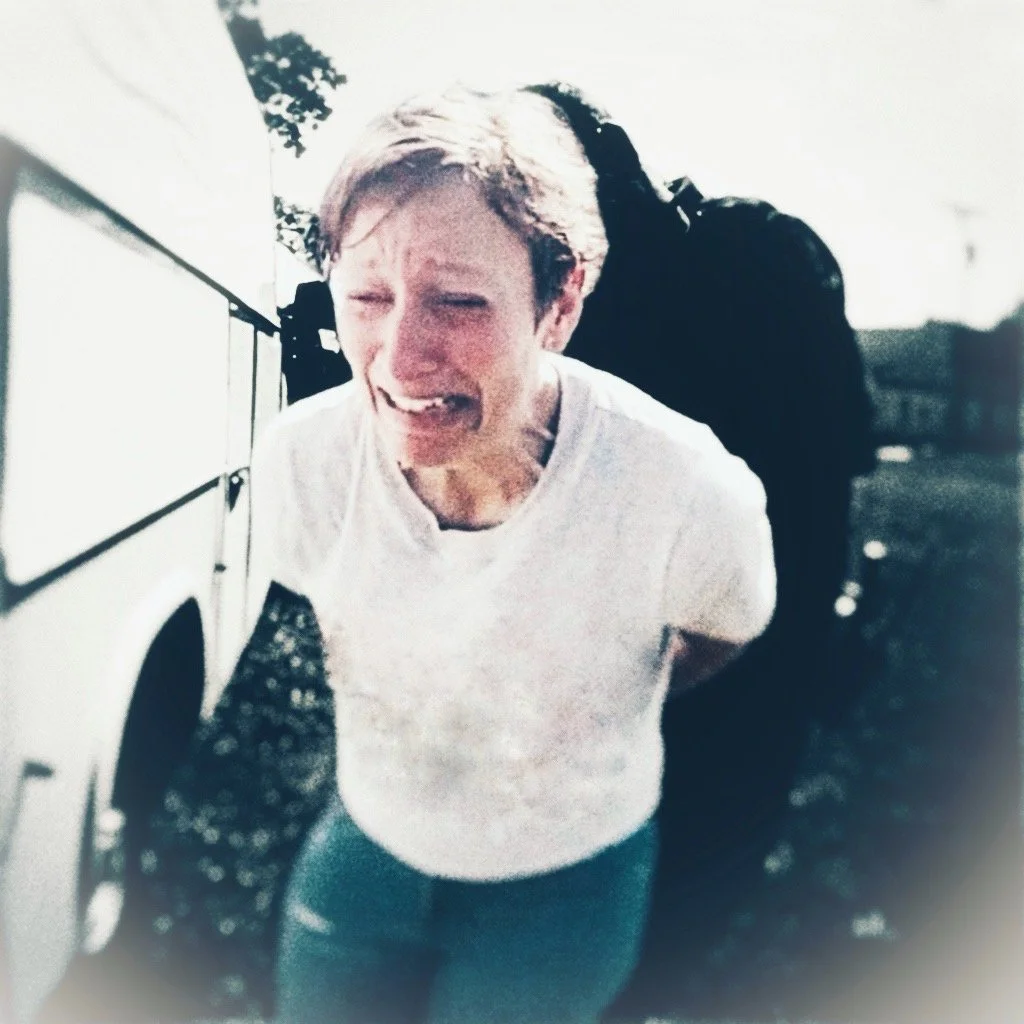 A distressed woman with short hair, wearing a white t-shirt and jeans, standing outdoors next to a vehicle, crying with her eyes closed.