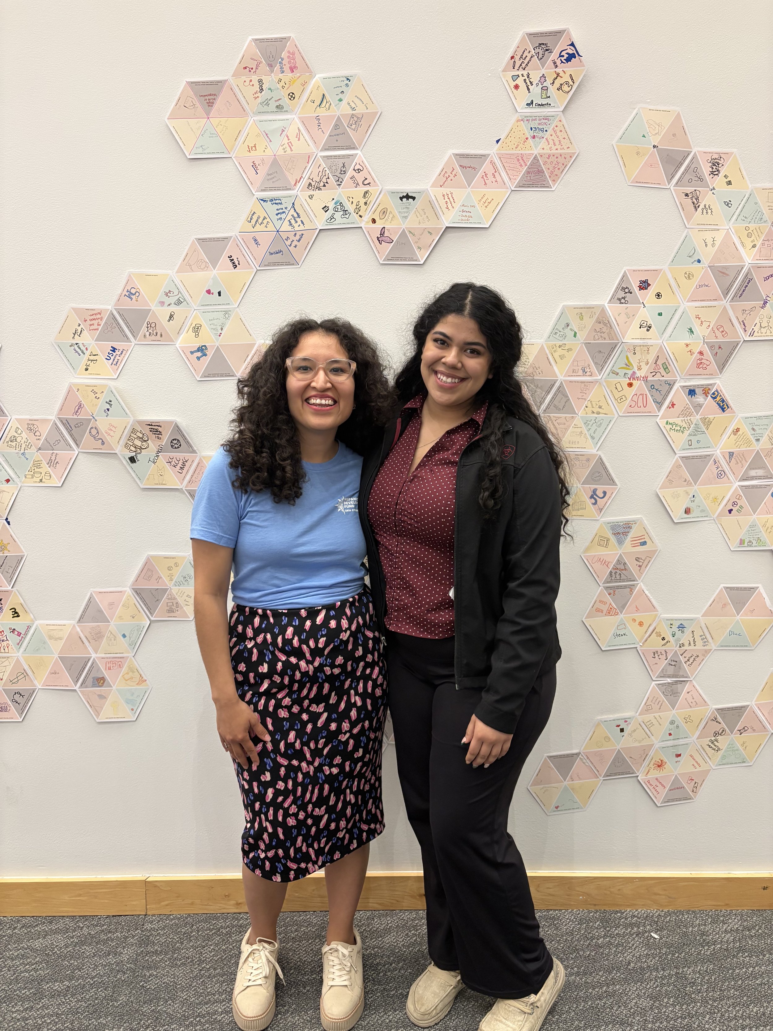Two women standing in front of a wall decorated with hexagonal paper tiles featuring drawings and writings, smiling at the camera.