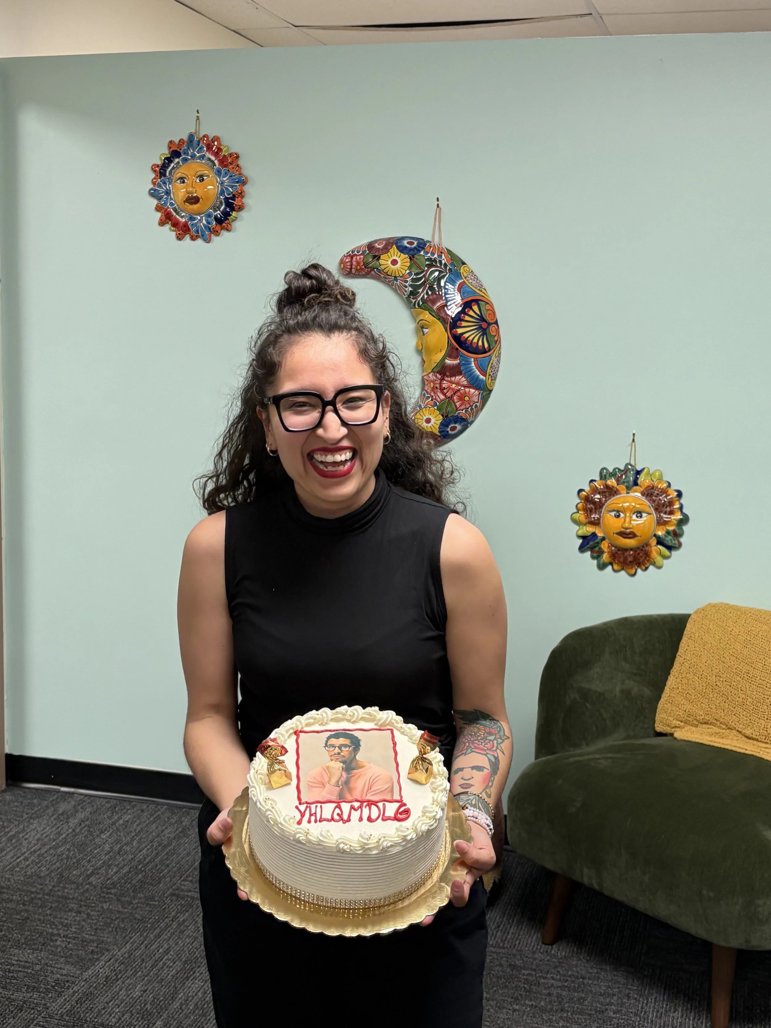 A woman with glasses and tattoos on her arm is smiling and holding a birthday cake. The cake features a framed picture of a man and a woman with the word 'YHLAMDLG' written in red icing. The woman is standing in a room with light green walls and deco