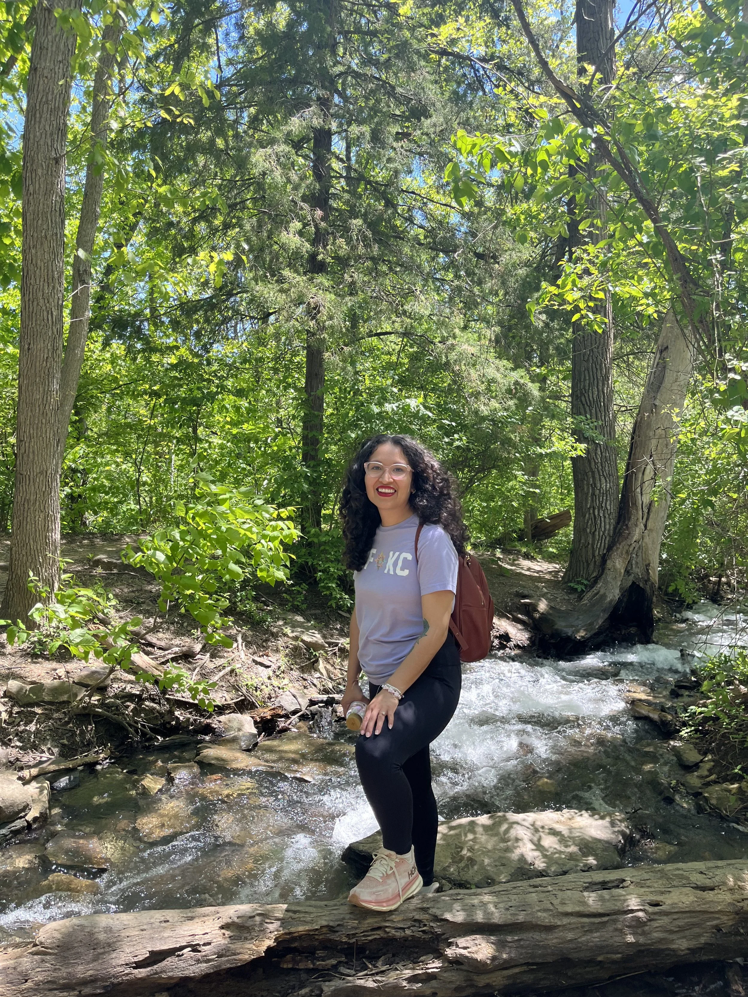 A woman with glasses and curly dark hair smiling while standing in a stream in a forest with tall trees and green leaves.