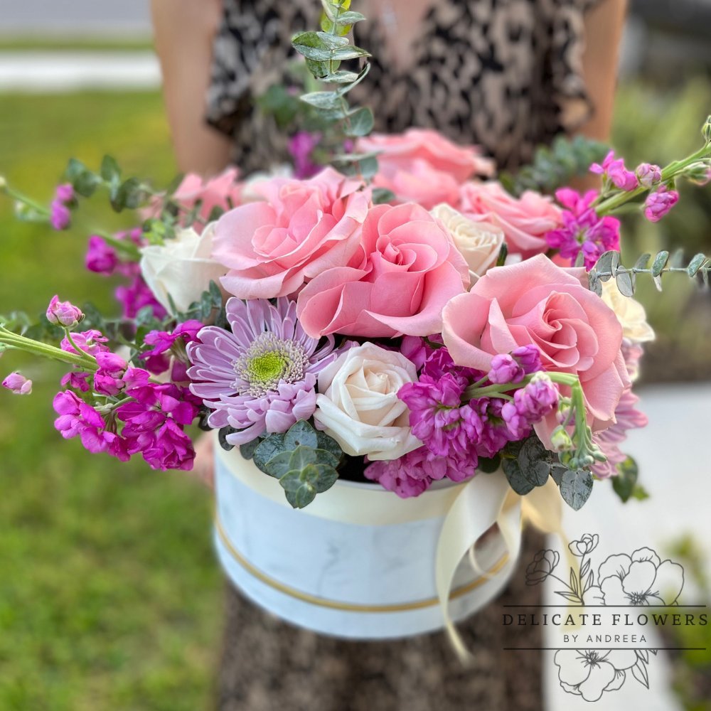 Bouquet of pink roses, purple daisies, and eucalyptus in a round floral box with logo.
