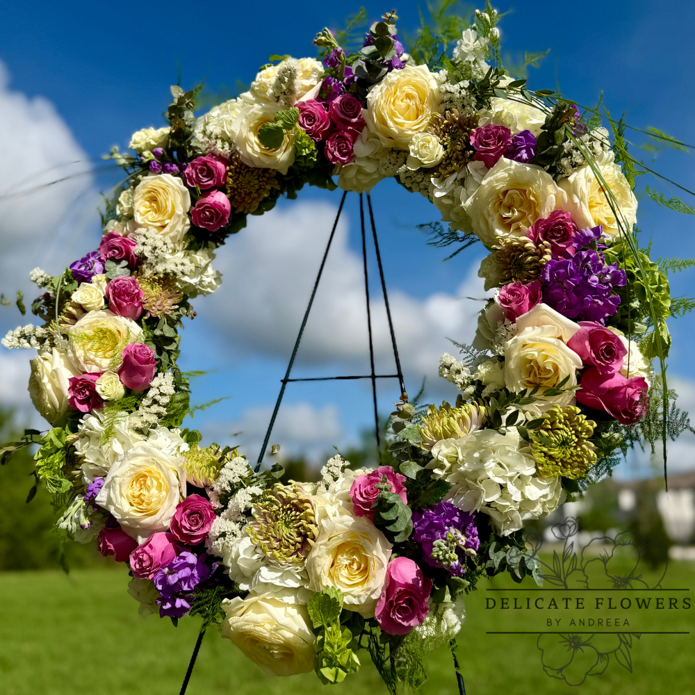 Standing celebration wreath with ivory roses, pink roses, purple seasonal flowers, and fresh greenery arranged in a natural garden style.