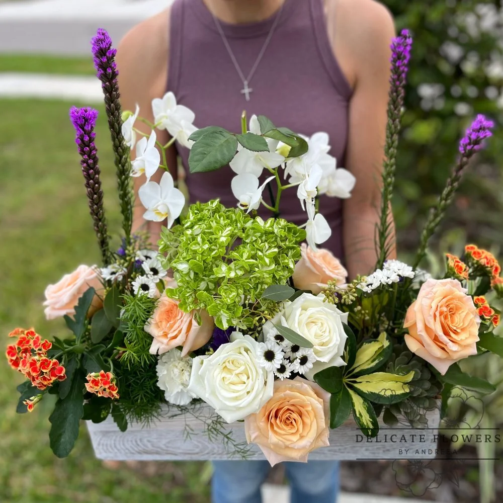a flower arrangement with roses, white orchids, and various other colorful flowers in a wooden box.