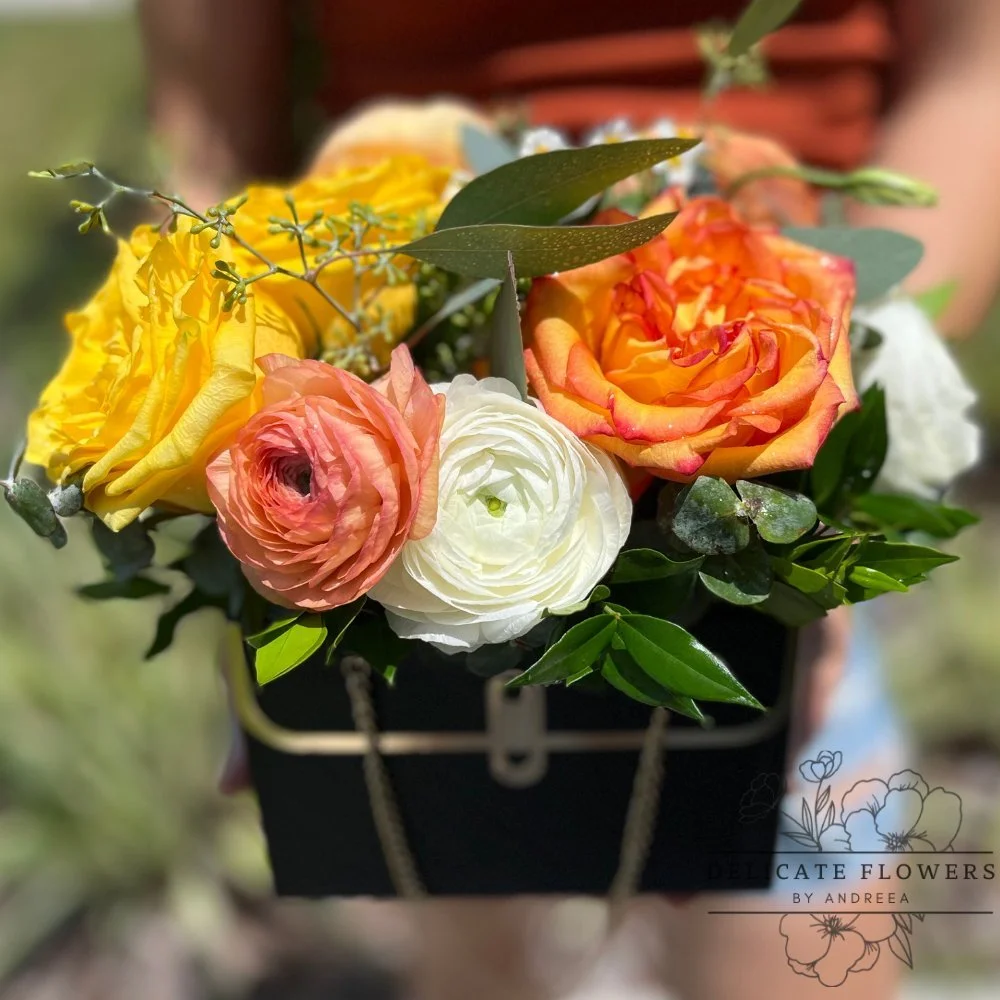 Black Beauty flower arrangement with yellow and orange roses, blush and ivory blooms, greenery, in a black container