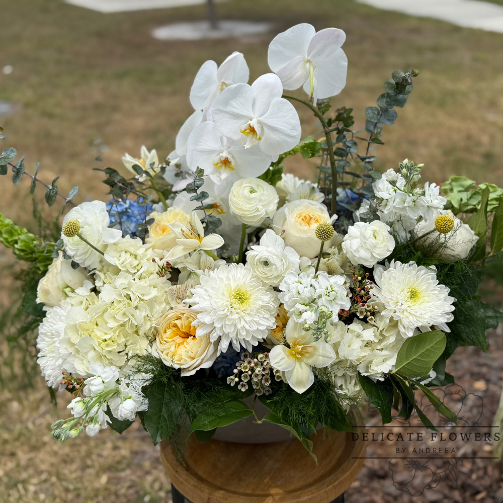 Elegant white floral arrangement with white orchids, roses, hydrangea, textured blooms, and lush greenery in a modern vessel.