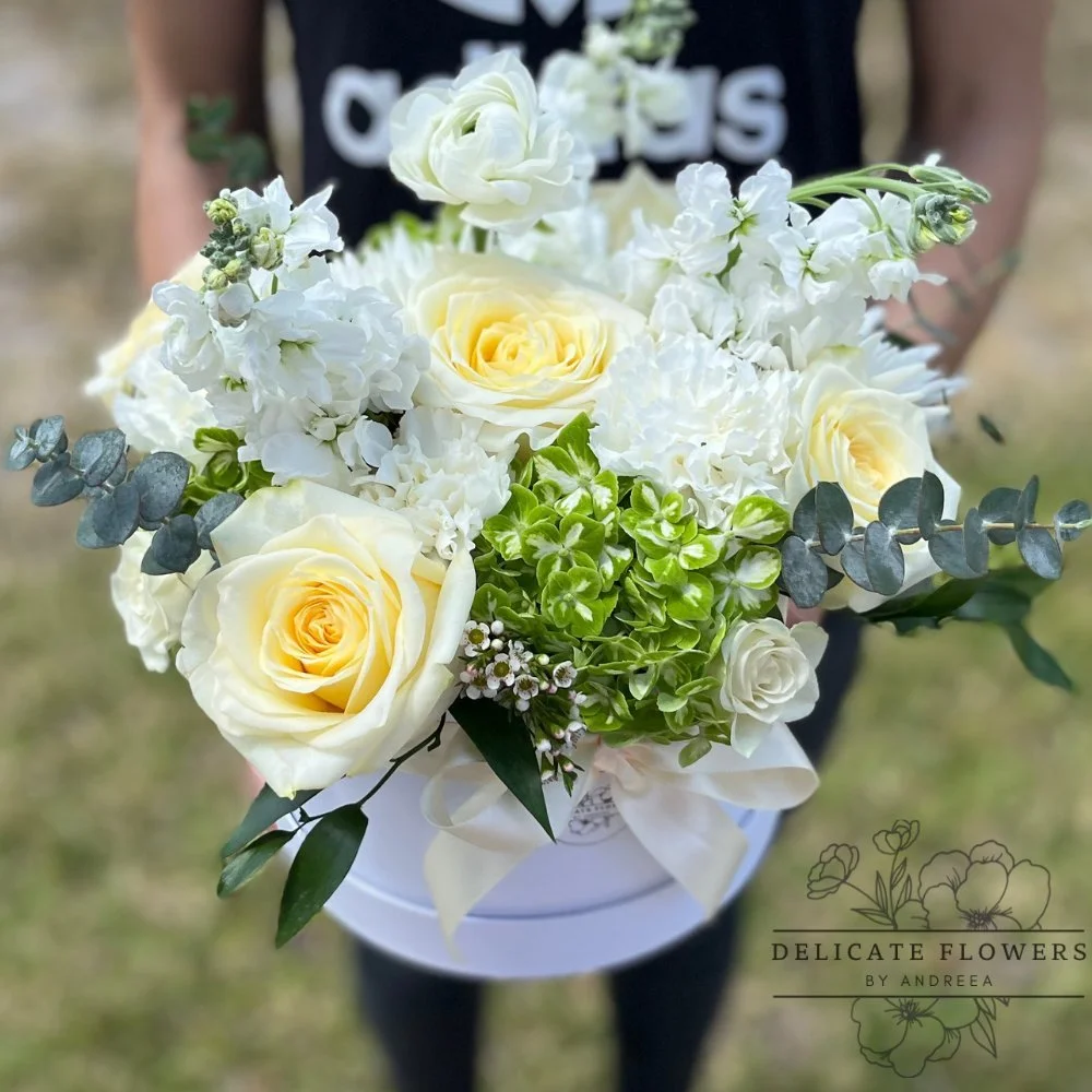 White floral hat box with ivory roses, ranunculus, green hydrangea, eucalyptus, and seasonal white blooms