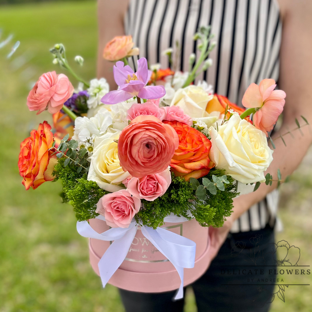 Lush floral arrangement featuring cream and coral roses, pink ranunculus, pastel accent blooms, and greenery, designed in a blush pink hat box with a white ribbon, held outdoors in natural light.