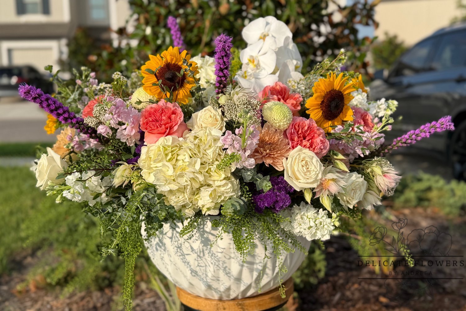 Garden Grandeur floral centerpiece featuring yellow sunflowers, white orchids, ivory roses, peach-pink flowers, hydrangeas, and purple accents in a white ceramic vase by Delicate Flowers by Andreea in Wesley Chapel, FL.