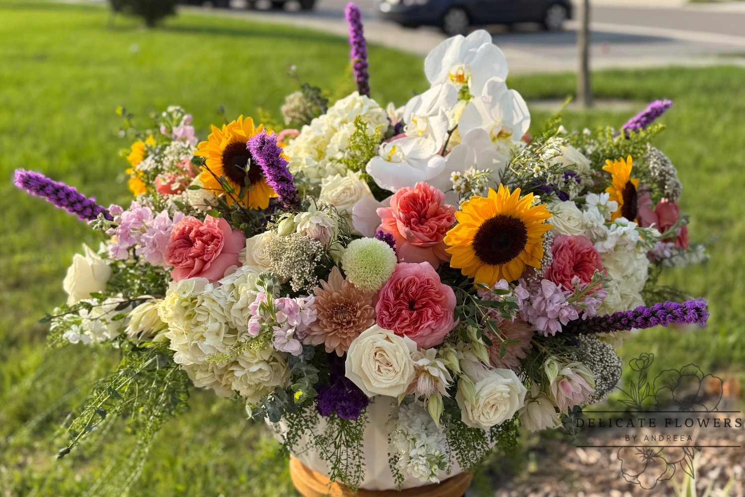 Luxury floral arrangement with white orchids, sunflowers, roses, hydrangeas, pink blooms, purple accents, and seasonal greenery