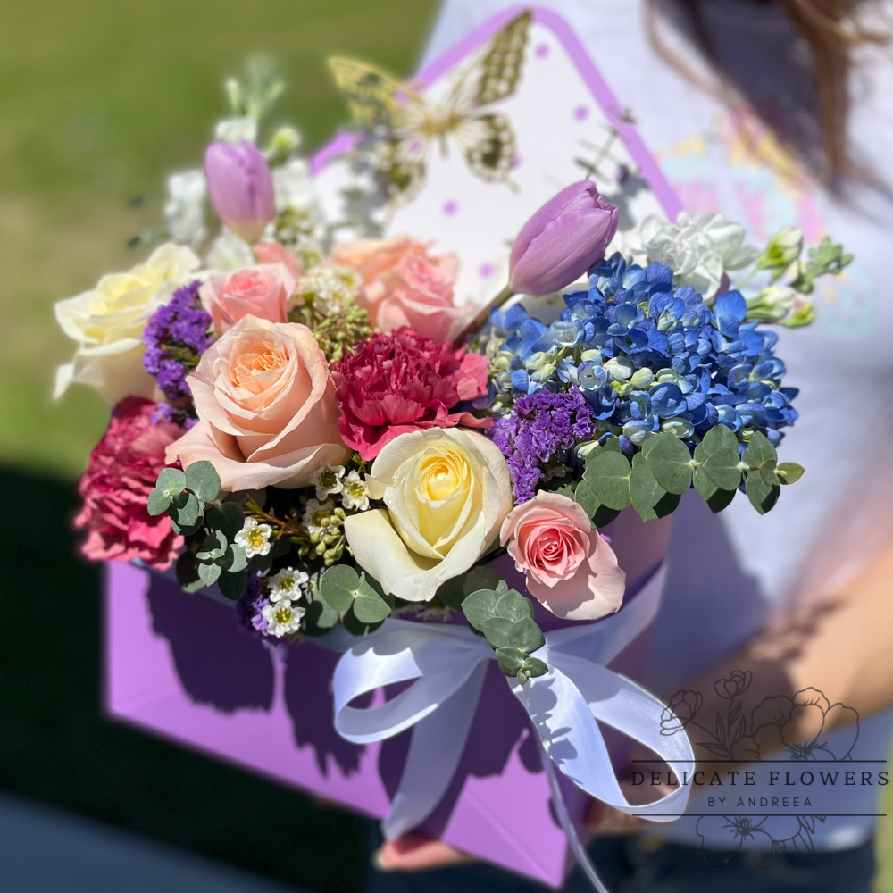 Elegant floral arrangement with pastel roses, pink tulips, blue hydrangea, purple accent flowers, and eucalyptus, styled in a lavender hat box with a white ribbon and decorative butterfly card, held outdoors.