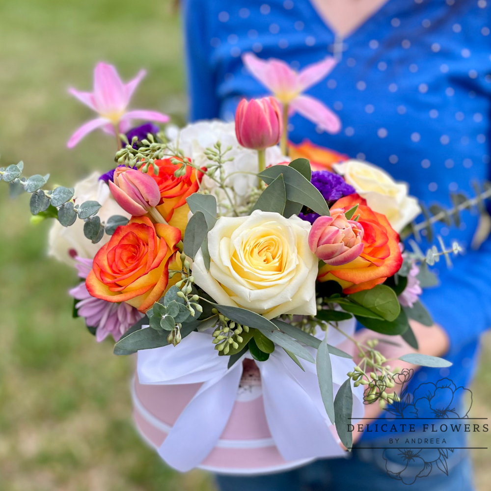 Colorful floral arrangement with cream and orange roses, pink tulips, purple accent flowers, eucalyptus, and greenery, arranged in a soft pink hat box with a white ribbon, held outdoors.