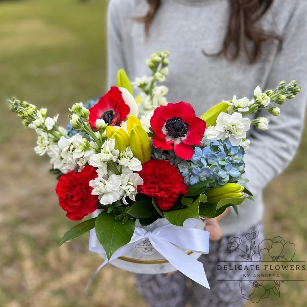 Colorful hat box arrangement with red anemones, red carnations, blue hydrangea, yellow lilies, and white stock