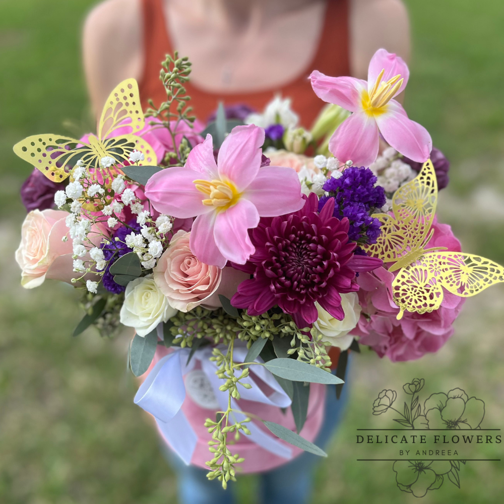 Handcrafted bouquet featuring pink lilies, blush and white roses, purple chrysanthemum, baby’s breath, and greenery, accented with decorative gold butterfly details and wrapped with a soft ribbon, held outdoors.