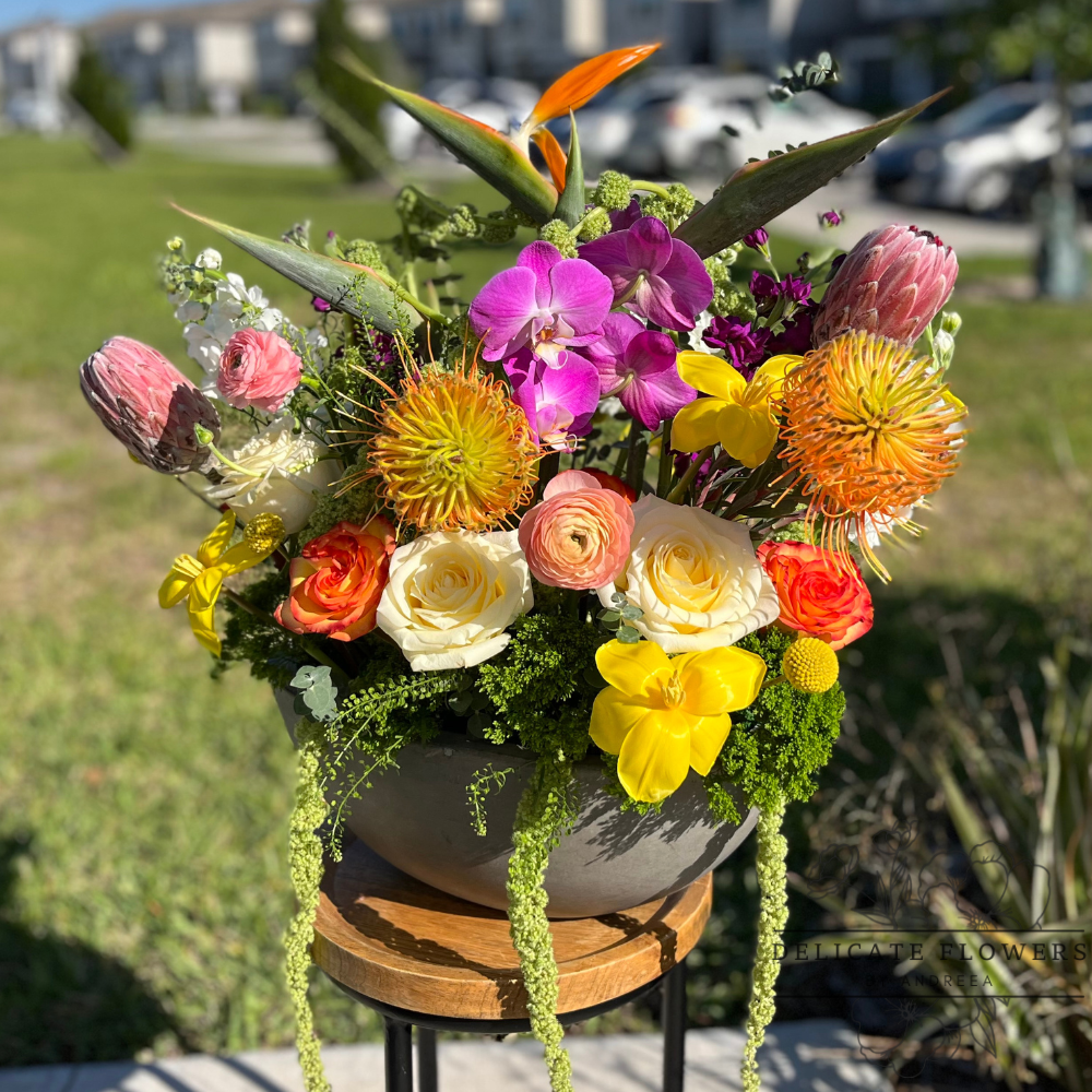 Bold, modern floral arrangement featuring bird of paradise, pink orchids, cream and orange roses, yellow accent blooms, pincushion protea, and lush greenery, styled in a contemporary gray bowl on a wooden stand outdoors.