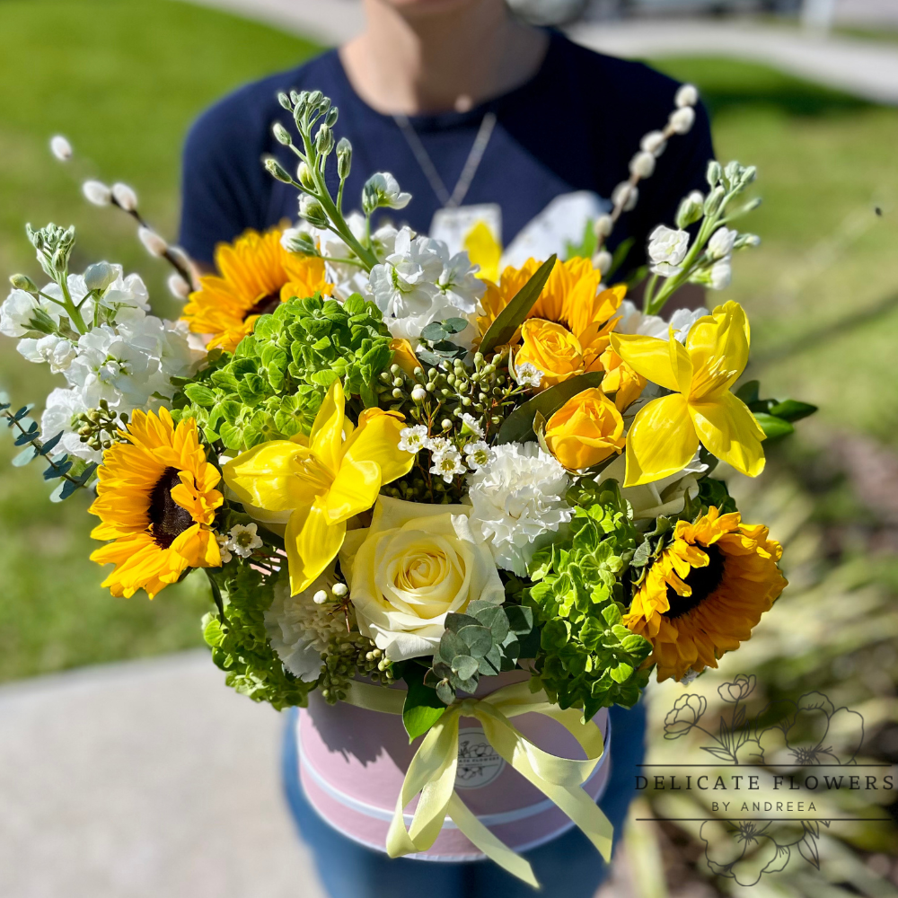 Bright floral arrangement with sunflowers, yellow tulips, white roses, green hydrangea, and white accent blooms, arranged in a pastel pink hat box with a ribbon, held outdoors in natural sunlight.