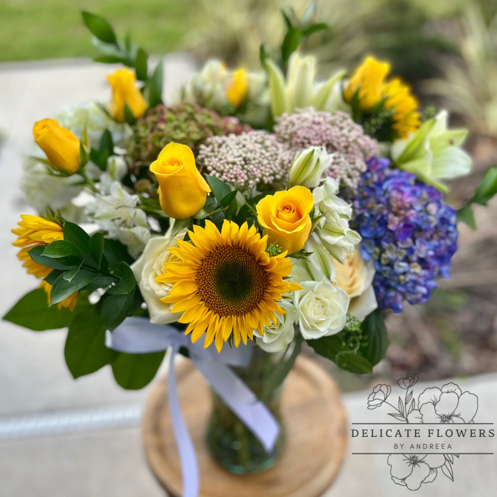 Fresh floral bouquet featuring sunflowers, yellow and white roses, blue hydrangea, lilies, and mixed greenery, arranged in a clear glass vase with a white ribbon, displayed outdoors.