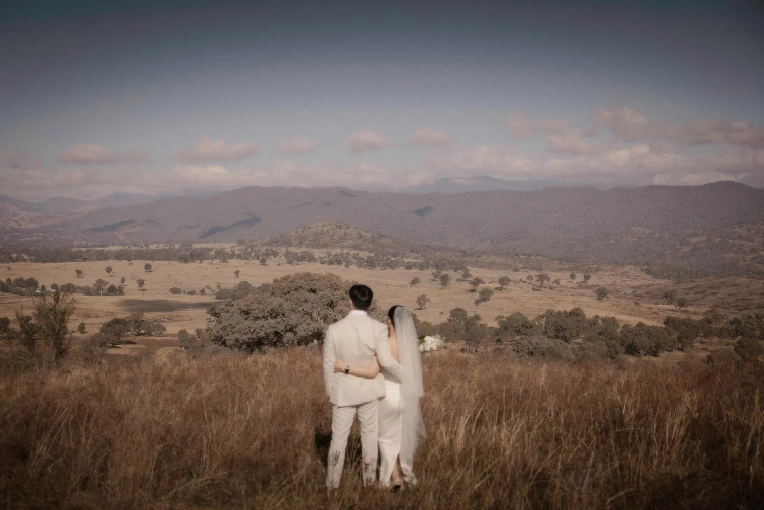 A bride and groom in wedding attire walking in a field with mountains in the background, during their adventure elopement wedding in Canberra by Wander & Be.