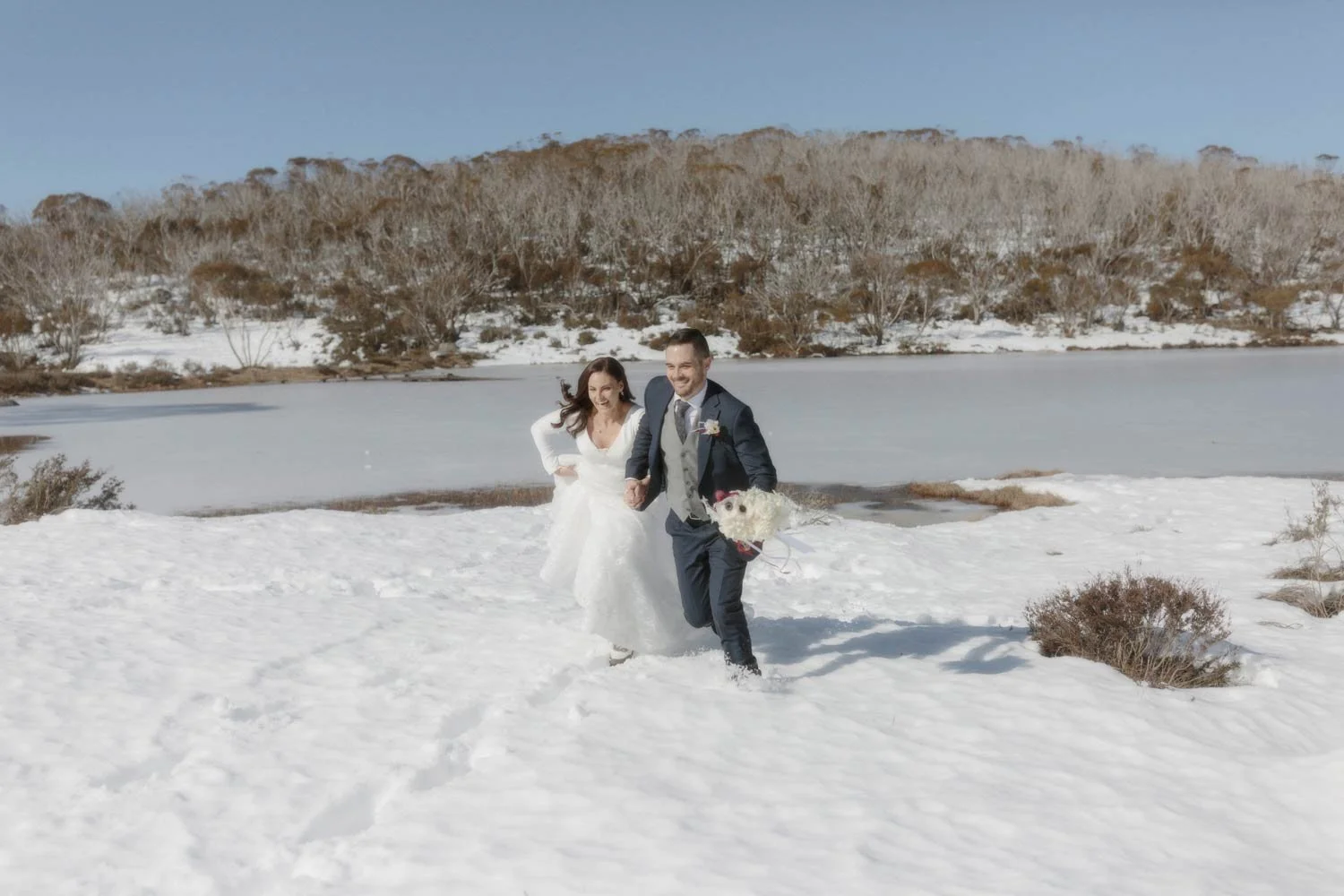 A bride and groom walking in snow near a frozen lake in the Snowy Mountains NSW holding hands and smiling, with trees and a clear blue sky in the background, during their adventure elopement wedding by Wander & Be.