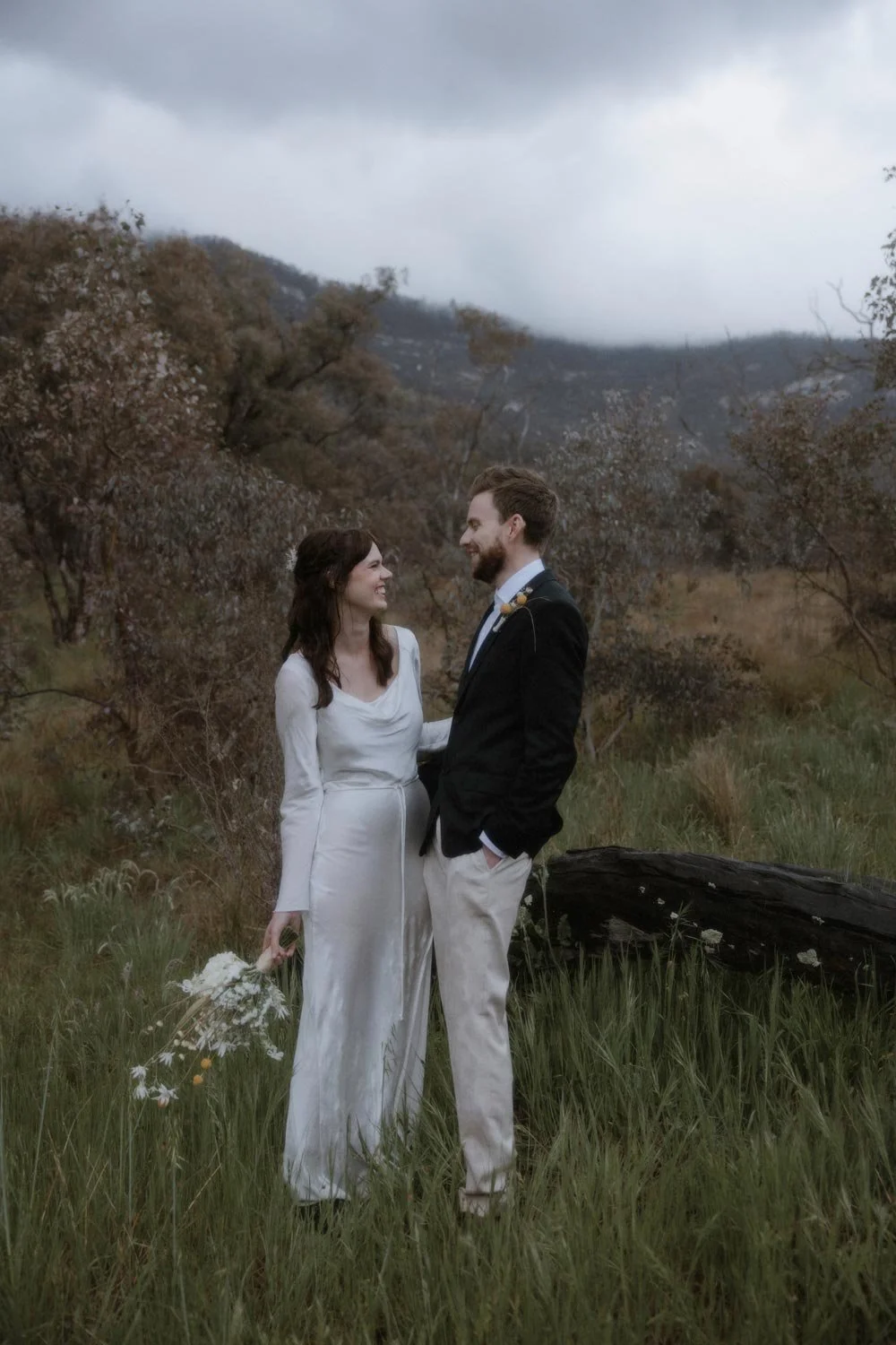 A couple in wedding attire standing in a grassy field with trees and mountains in the background, sharing a moment and smiling at each other, during their adventure elopement wedding in Canberra by Wander & Be.