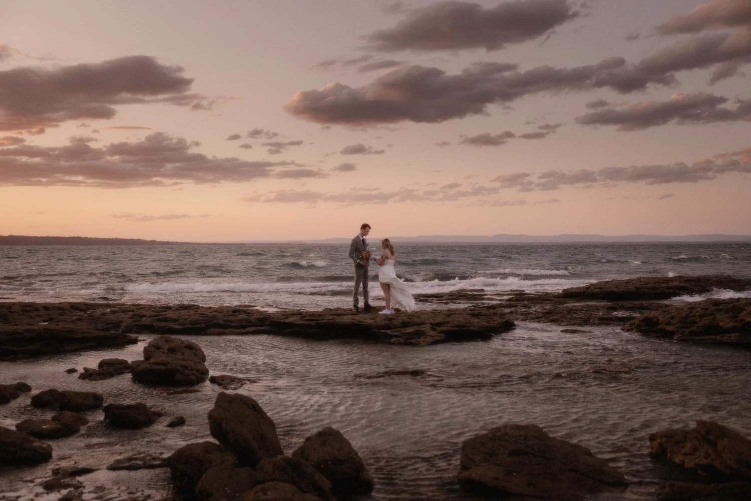 A couple dressed in wedding attire standing on a rocky shoreline during sunset, holding hands and facing each other by the ocean, during their adventure elopement wedding by Wander & Be.