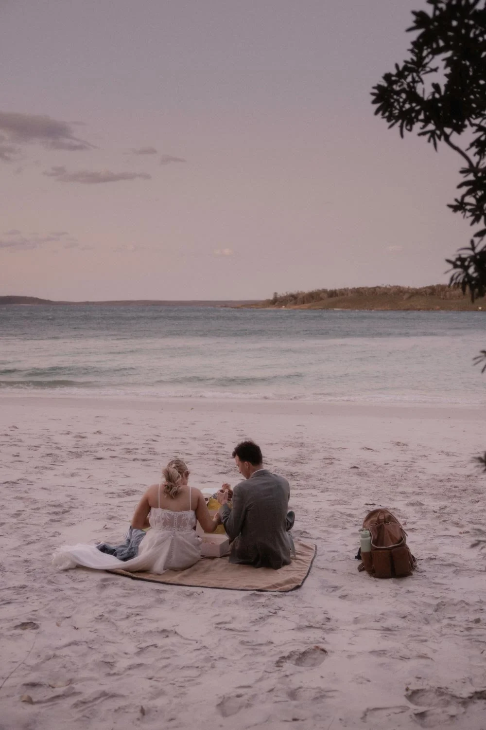 A couple having a picnic on the beach at sunset, sitting on a blanket with a backpack nearby, near the ocean with a small island or landmass in the background, during their South Coast NSW adventure elopement wedding by Wander & Be.