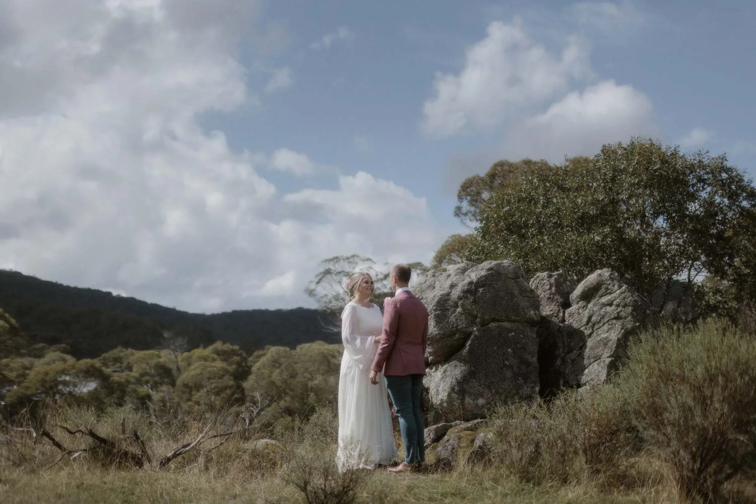 A bride and groom stand outdoors on a grassy field with rocks and trees in the Snowy Mountains NSW during spring, facing each other holding hands, during their elopement wedding ceremony by Wander & Be.