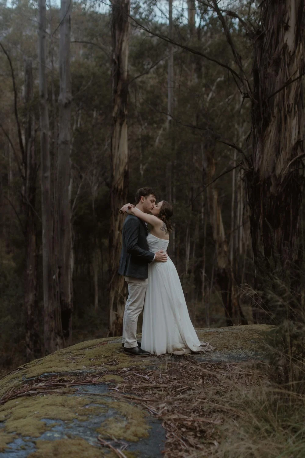 A couple in wedding attire sharing a kiss in a forest setting, with tall trees and moss-covered ground, during their adventure elopement wedding in Canberra by Wander & Be.
