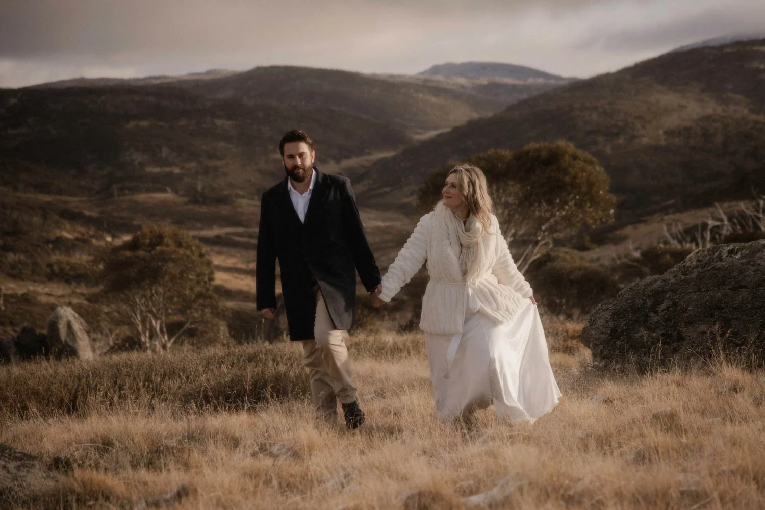 A man in a black coat and beige pants holding hands with a woman in a white dress and coat walking across an open field with hills and trees in the background, during their Snowy Mountains NSW adventure elopement wedding by Wander & Be.