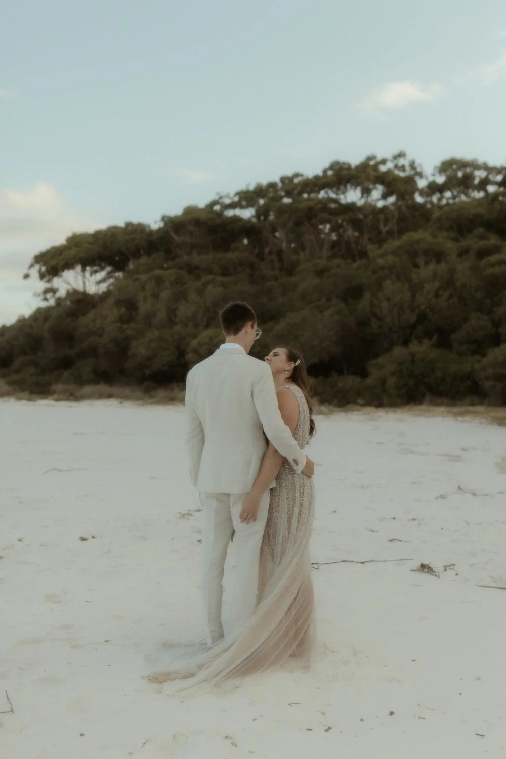 A bride and groom standing on a sandy beach, holding each other and facing each other, with a backdrop of trees and a blue sky, during their adventure elopement wedding by Wander & Be.