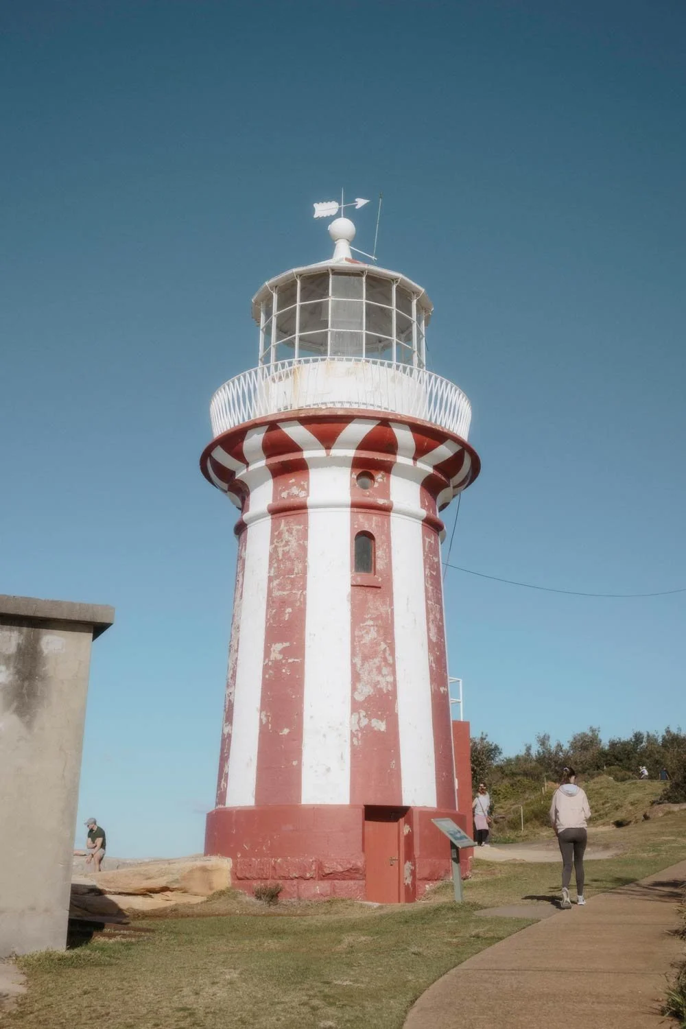 Red and white striped lighthouse in Sydney with a glass-enclosed top, situated outdoors on a clear day. There are a few people walking near it and a grassy area.