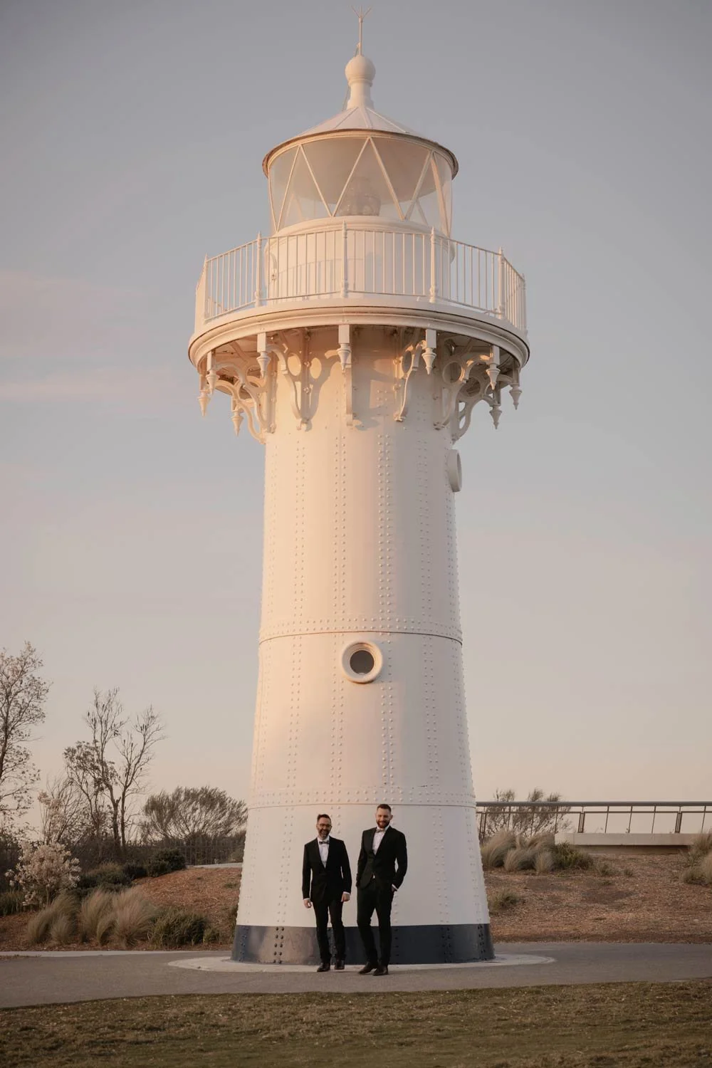 A white lighthouse with two men in tuxedos standing at its base at sunset, during their South Coast NSW adventure elopement wedding by Wander & Be.