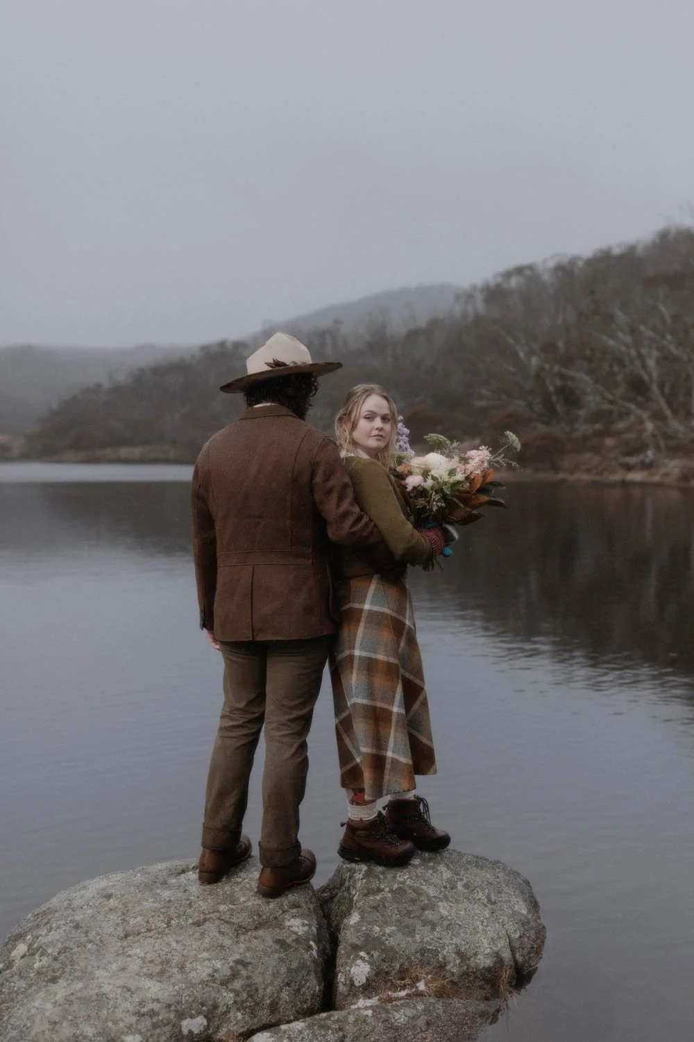 A man and woman standing on a large rock by a lake in autumn or winter, with the woman holding a bouquet of flowers, both dressed in vintage-style clothing during their adventure elopement wedding by Wander & Be.