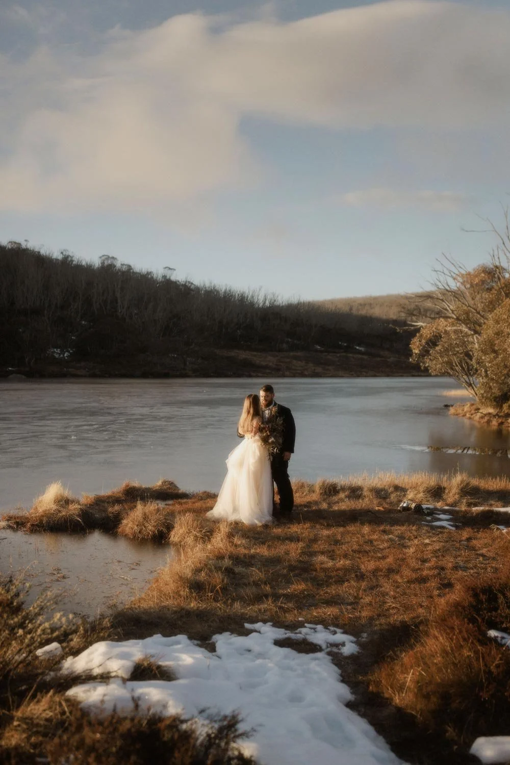 A bride and groom standing on a grassy bank beside a frozen lake in the Snowy Mountains NSW, embracing each other during sunset, with leafless trees and a cloudy sky in the background, during their adventure elopement wedding by Wander & Be.