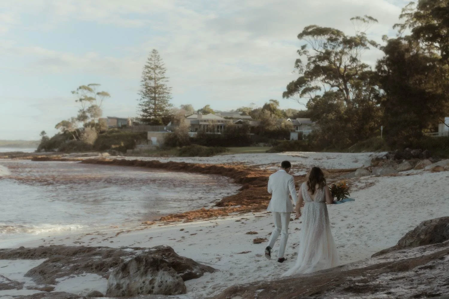 A couple dressed in wedding attire walking along a sandy beach, holding hands, with houses and trees in the background at their adventure elopement wedding by Wander & Be.