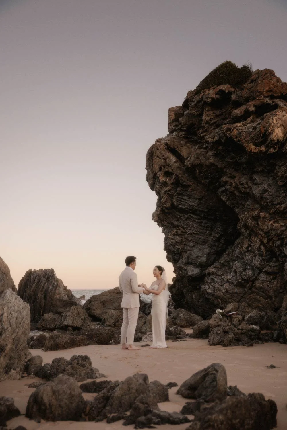 A couple dressed in wedding attire stands holding hands on a sandy beach, surrounded by large rocks and a coastal landscape during sunset, during their adventure elopement wedding in Canberra by Wander & Be.