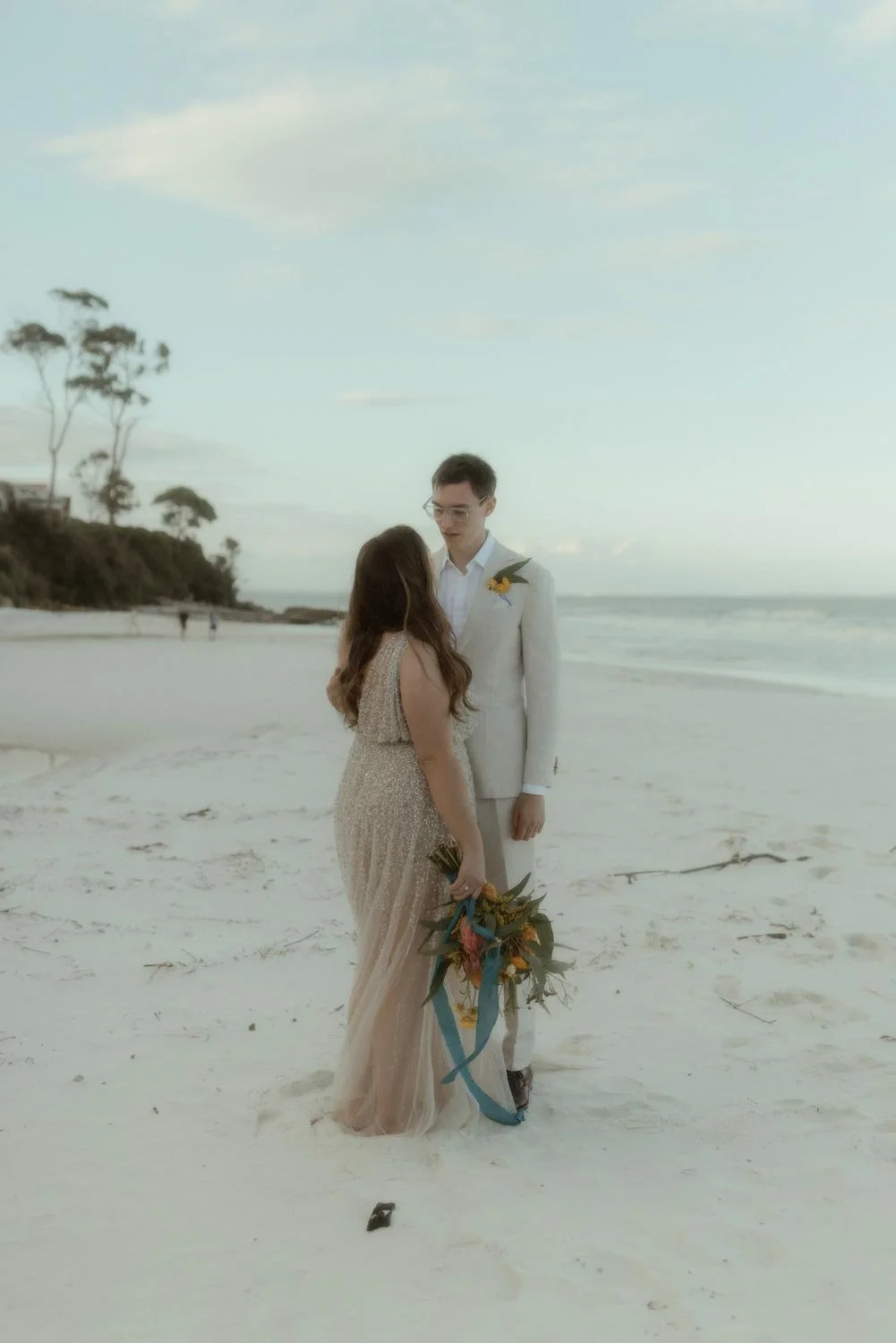A bride and groom standing on the beach, facing each other, holding a bouquet, with the ocean and sky in the background during their adventure elopement wedding by Wander & Be.
