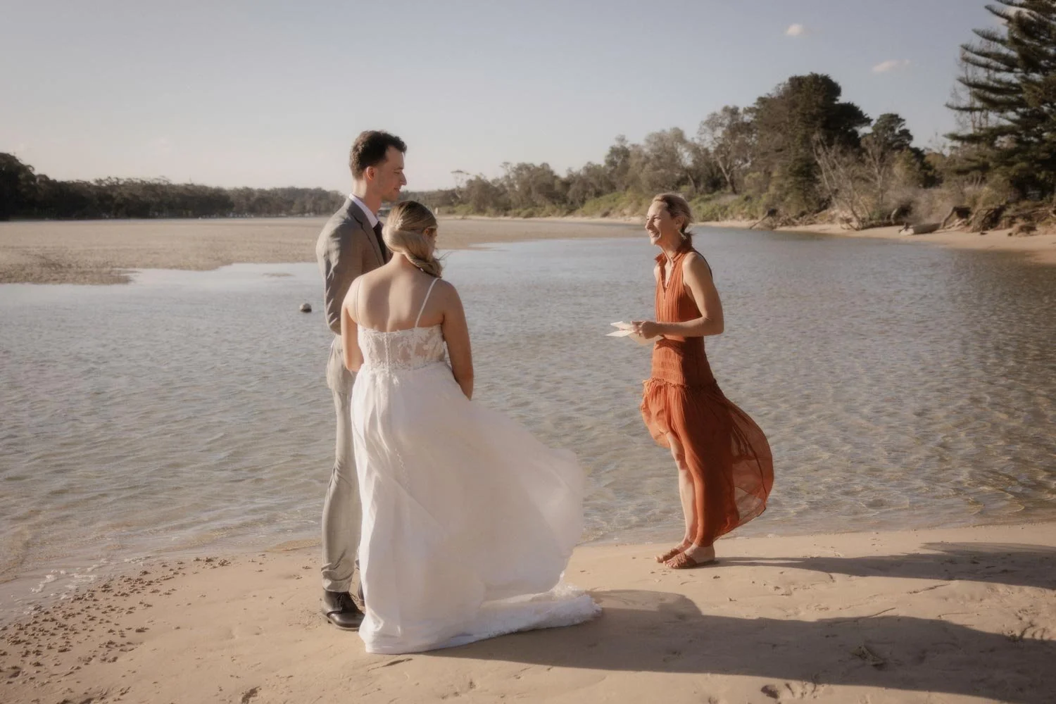 A couple getting married on the beach by Jacqua Celebrant officiating, near a river and trees under a clear sky for their South Coast NSW adventure elopement wedding by Wander & Be.