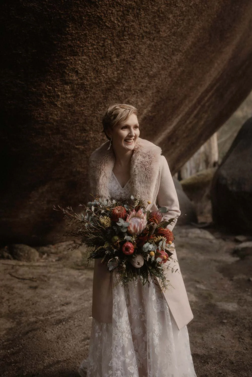 A smiling bride in a wedding dress and coat holding a bouquet of flowers, standing outdoors near large rocks, during her adventure elopement wedding in Canberra by Wander & Be.