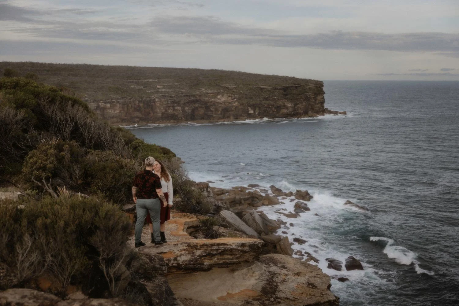 Two women standing on a rocky cliffside overlooking the ocean, with a large cliff in the background, during their adventure elopement wedding by Wander & Be in Sydney.