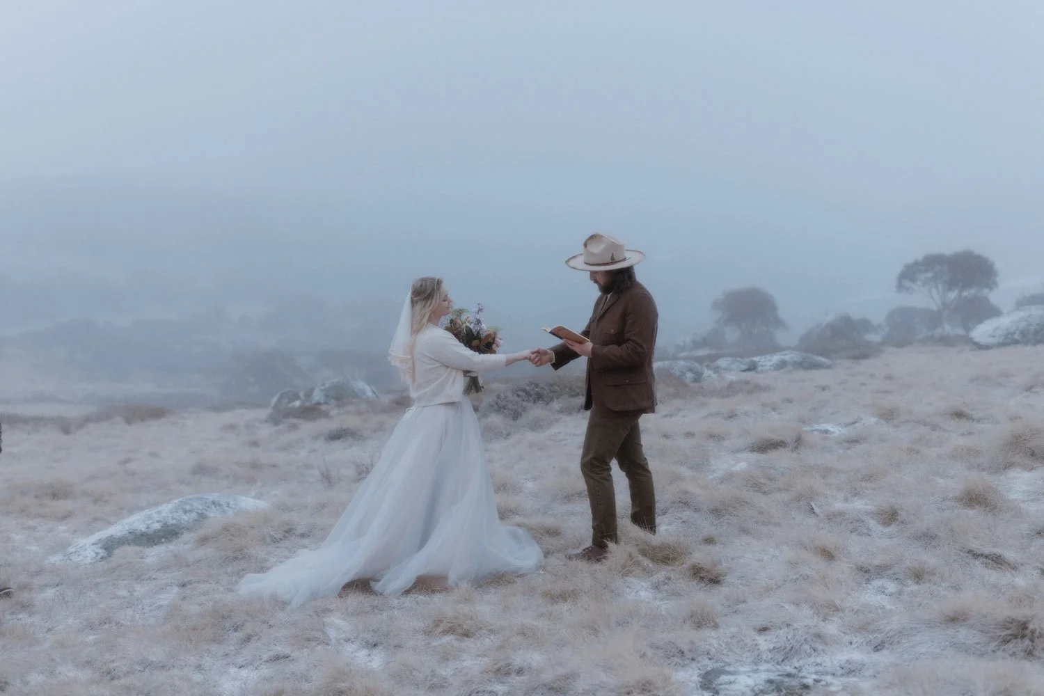 A bride in a white dress and veil holding her grooms hand, who's in a brown suit and wide-brimmed hat, outdoors on a foggy, rocky, Snowy Mountains NSW terrain, holding a bouquet of flowers, during their adventure elopement wedding by Wander & Be.