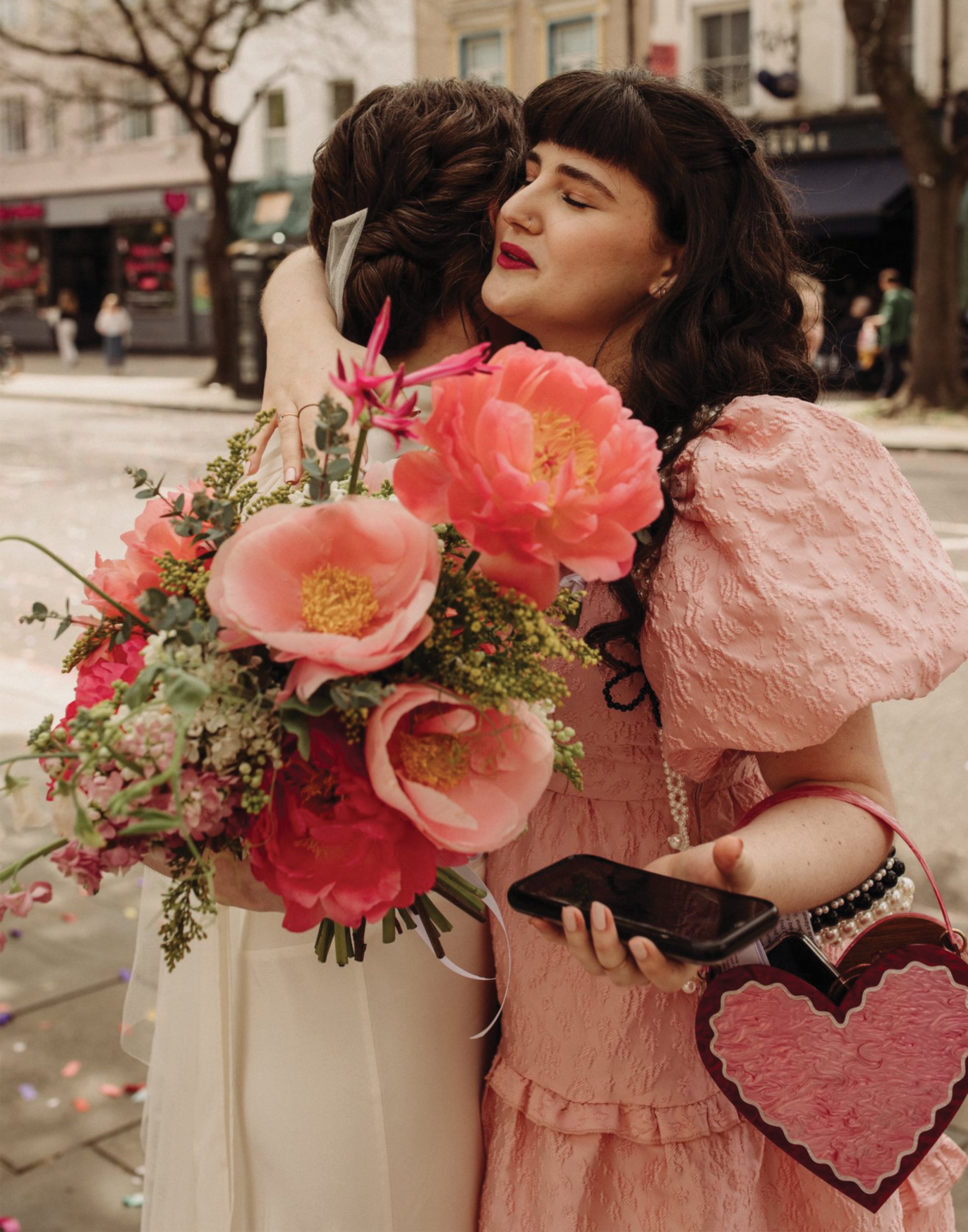 Two women hug, one in a wedding dress holding a bouquet of pink flowers, the other in a pink lace dress with dark curly hair, holding a smartphone. They are outdoors on a city street lined with trees and buildings.