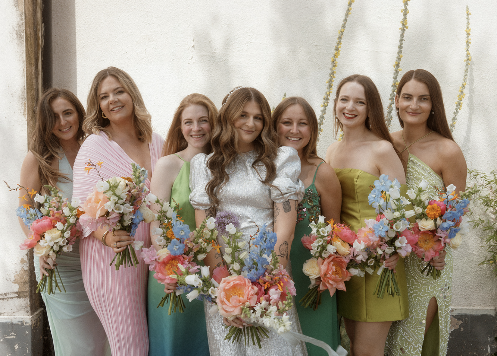 A group of eight women dressed in colorful dresses, holding bouquets of flowers, standing together against a plain wall with some decorative strands hanging from above.