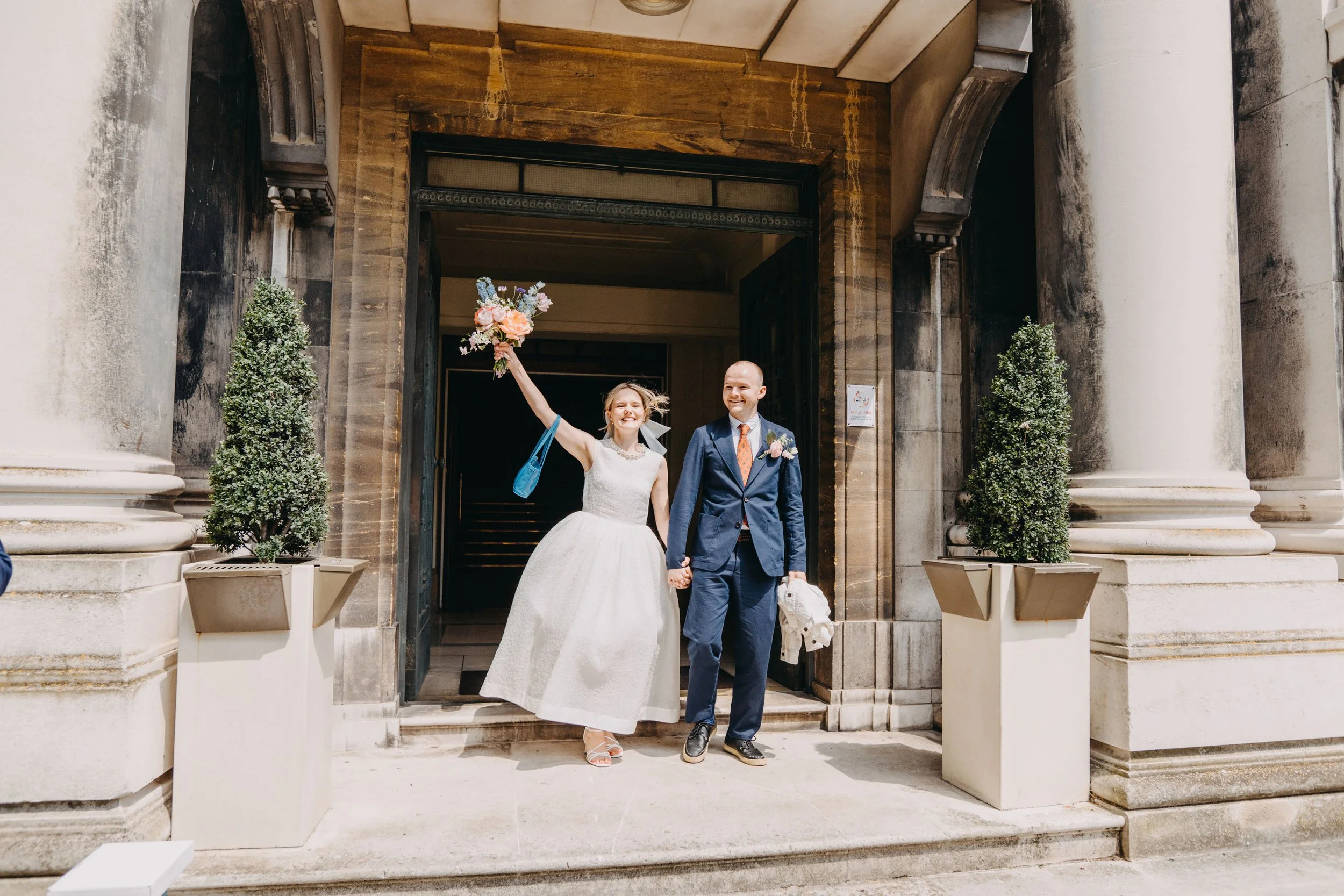 A newlywed couple, holding hands, walking out of a building with columns, the bride is smiling and holding a bouquet, the groom is smiling and holding a hat, both dressed in wedding attire.