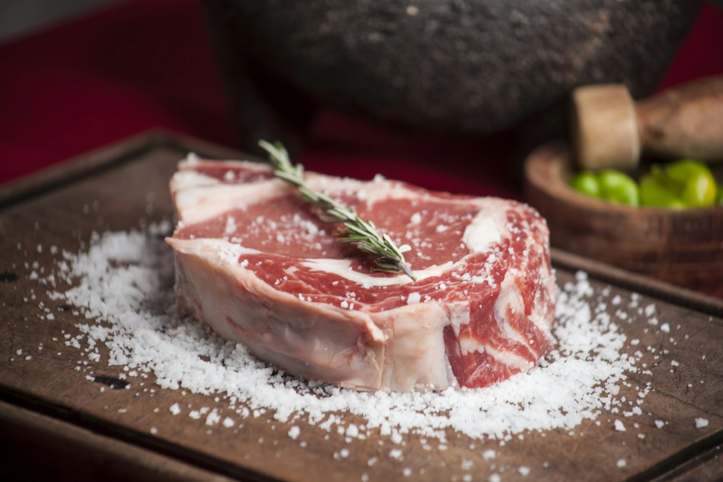 Raw bone-in chop seasoned with salt, garnished with a sprig of rosemary, on a wooden surface sprinkled with salt, with a bowl of green vegetables and a pepper grinder in the background.