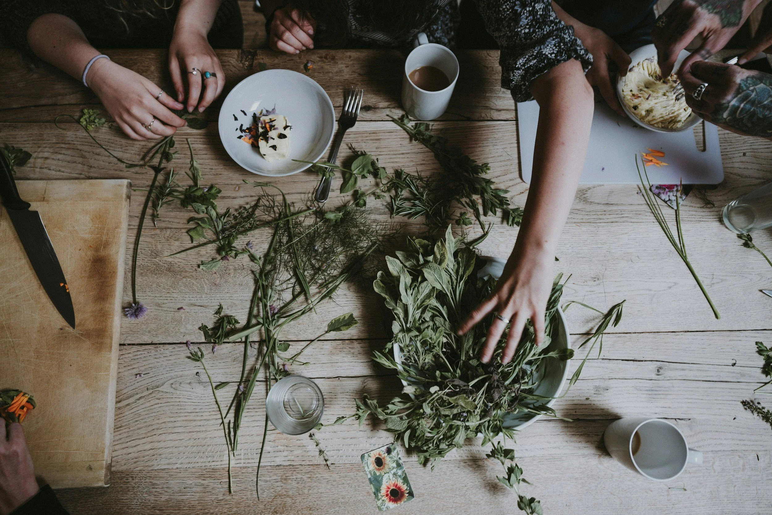 People working on a floral arrangement at a wooden table with various flowers, herbs, and tools, including a knife, chopping board, and cups of coffee.