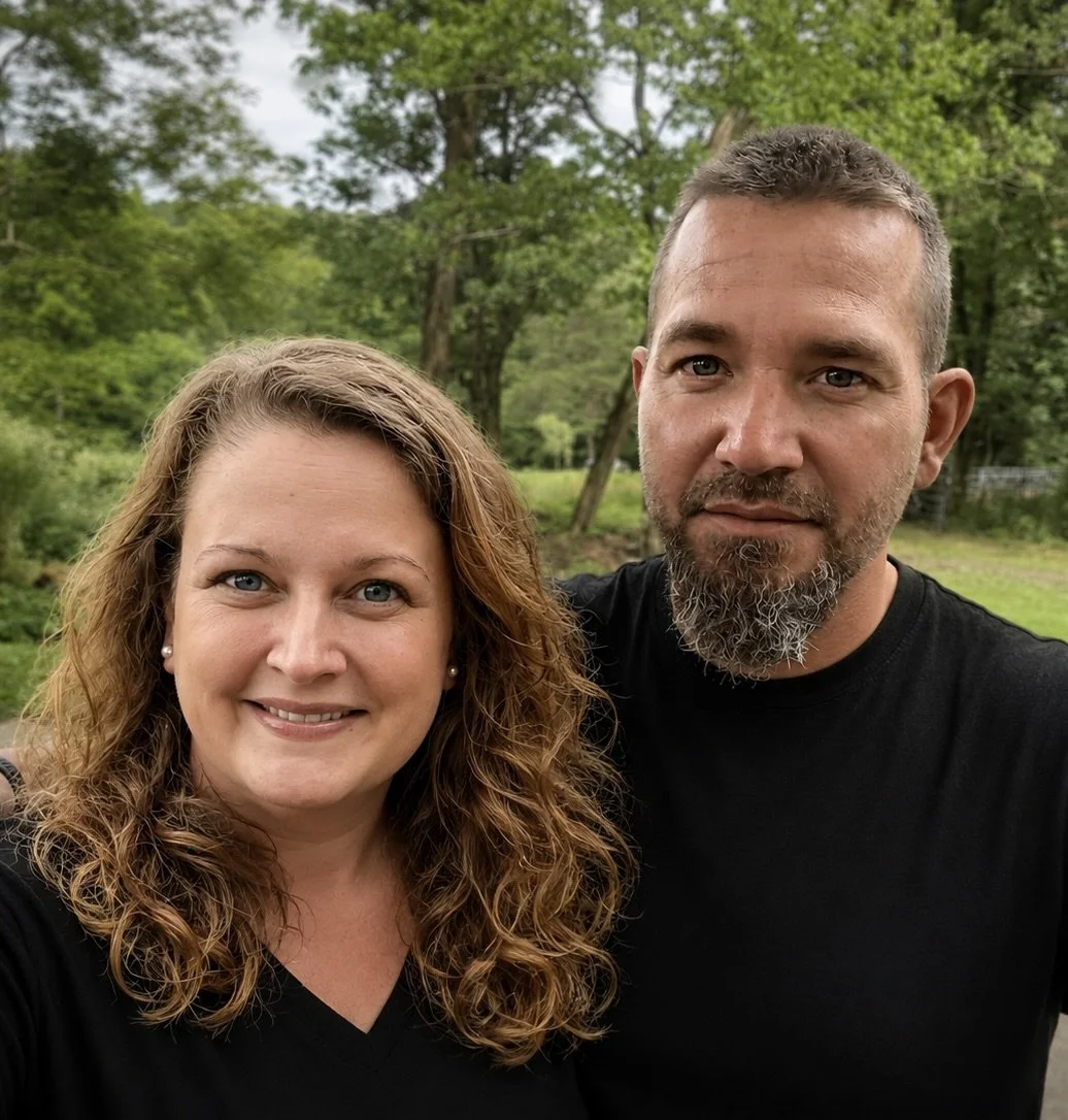 A smiling woman with curly hair and a bearded man standing outdoors surrounded by green trees.