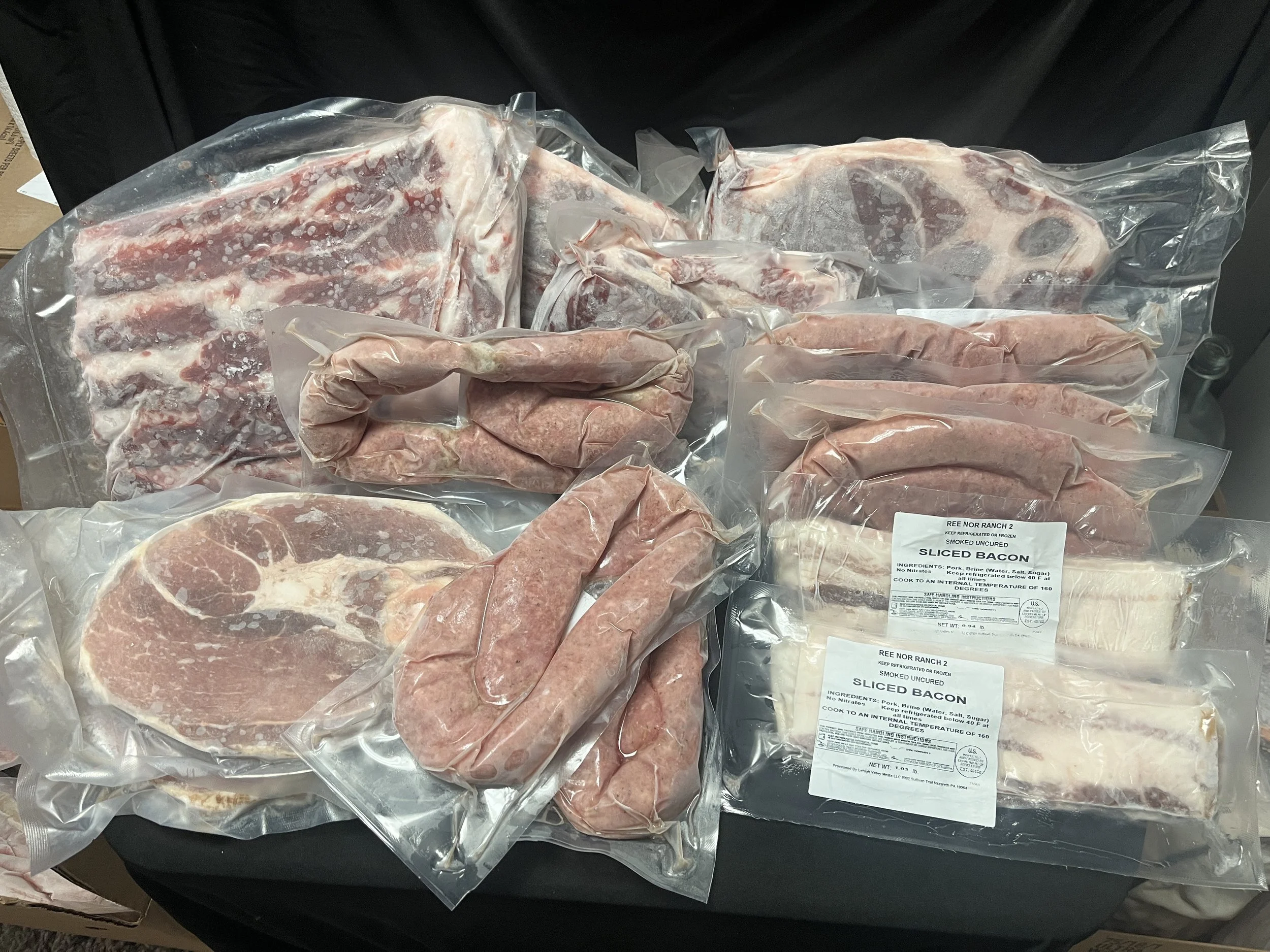 Assorted vacuum-sealed raw meat cuts including bacon, sausage links, steak, and ribs on a black surface.