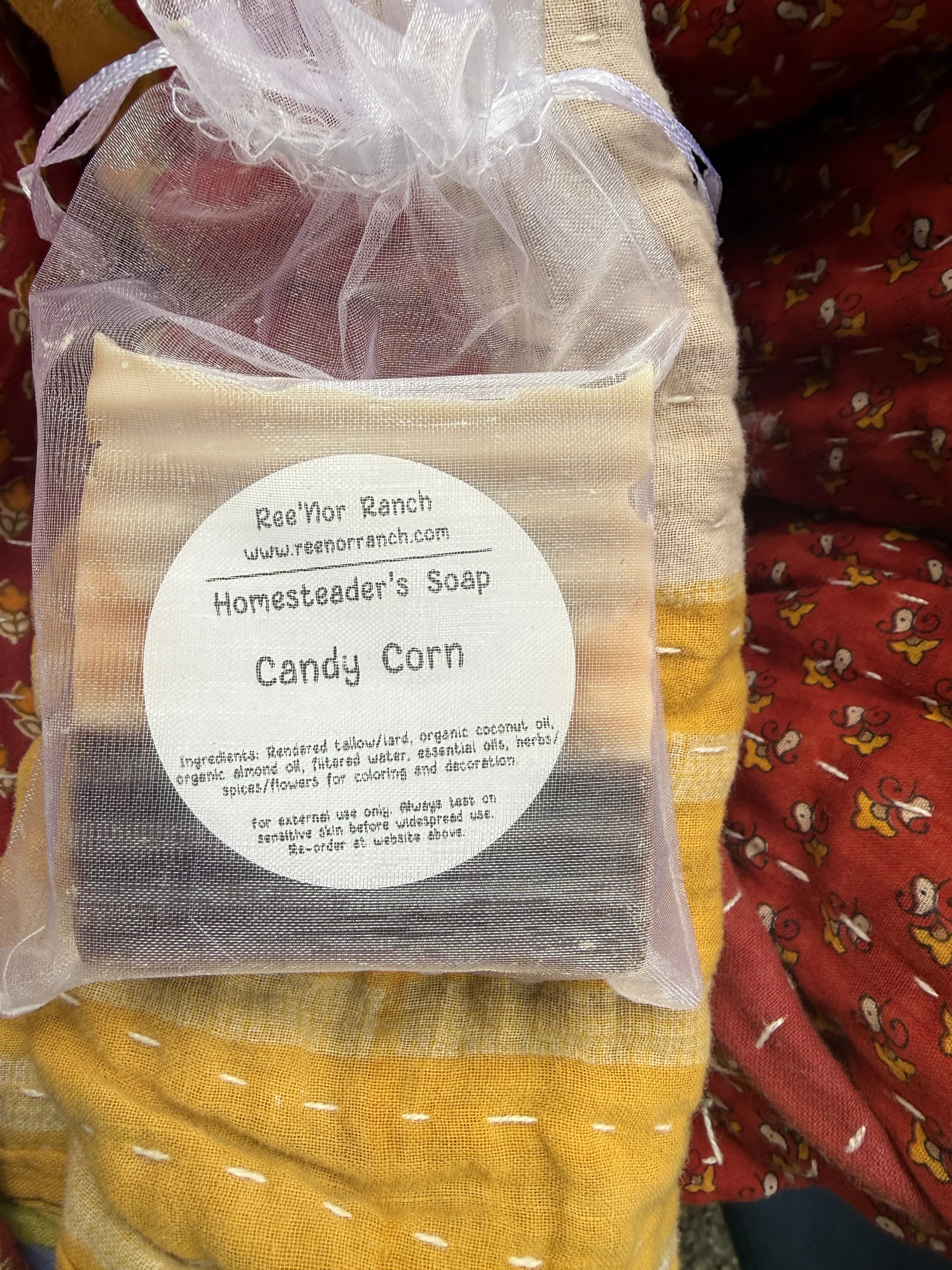 A clear plastic bag containing soap labeled 'Candy Corn' from Re'Nor Ranch, placed on a yellow cloth with white stitching, with a red cloth featuring a duck pattern in the background.
