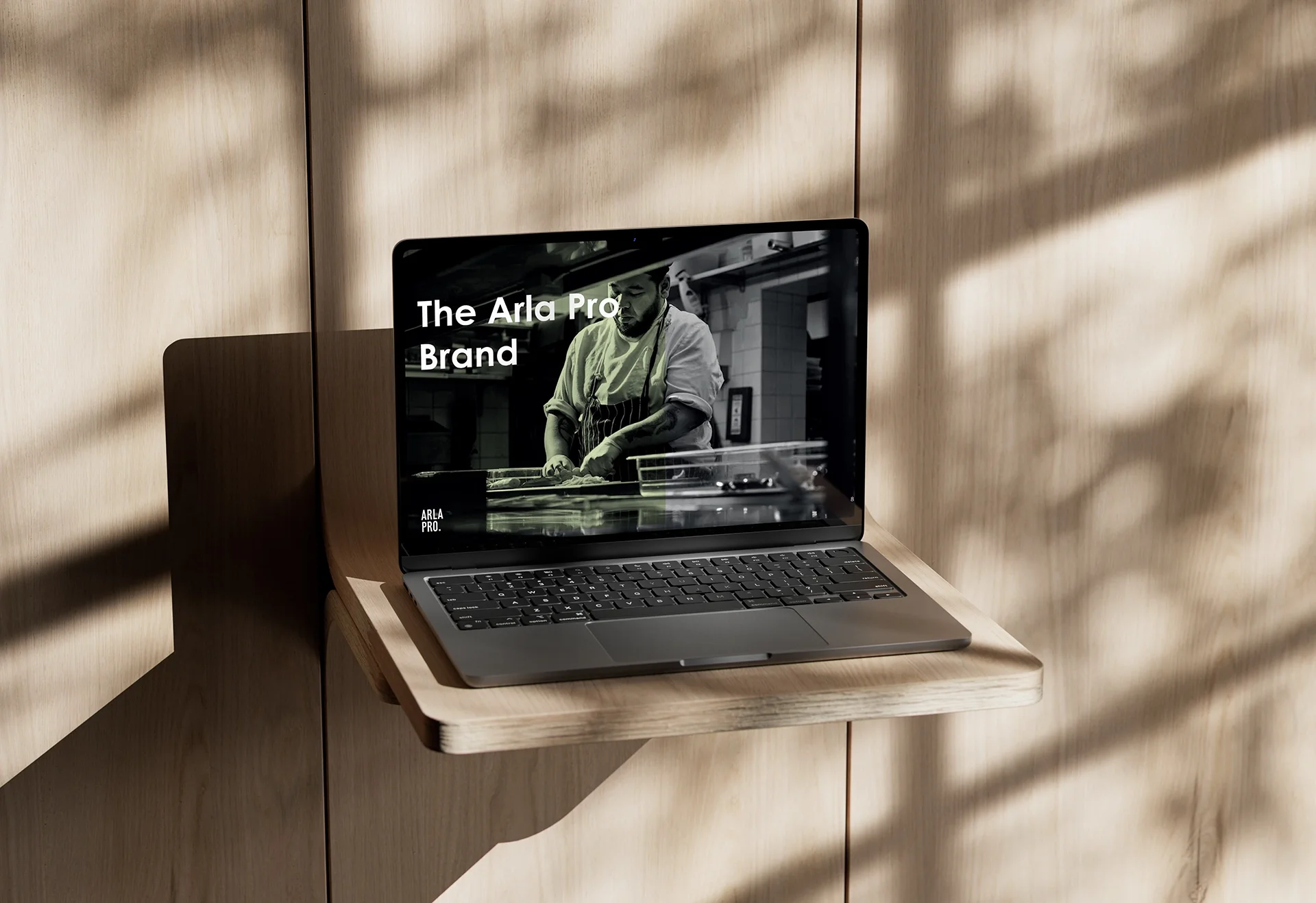A laptop on a wooden wall-mounted shelf displaying a chef working in a kitchen with the text "The Arla Pro Brand" on the screen.