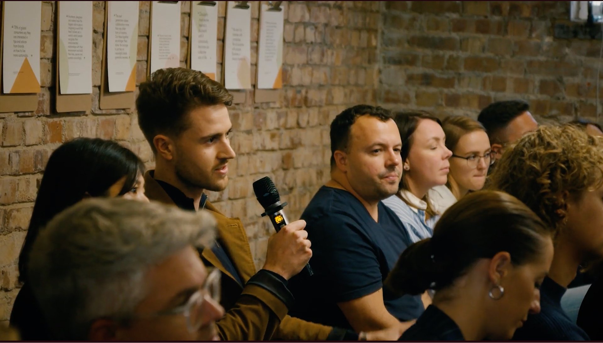 People sitting in a row attentively listening at an event, a man holding a microphone, brick wall background with framed documents.